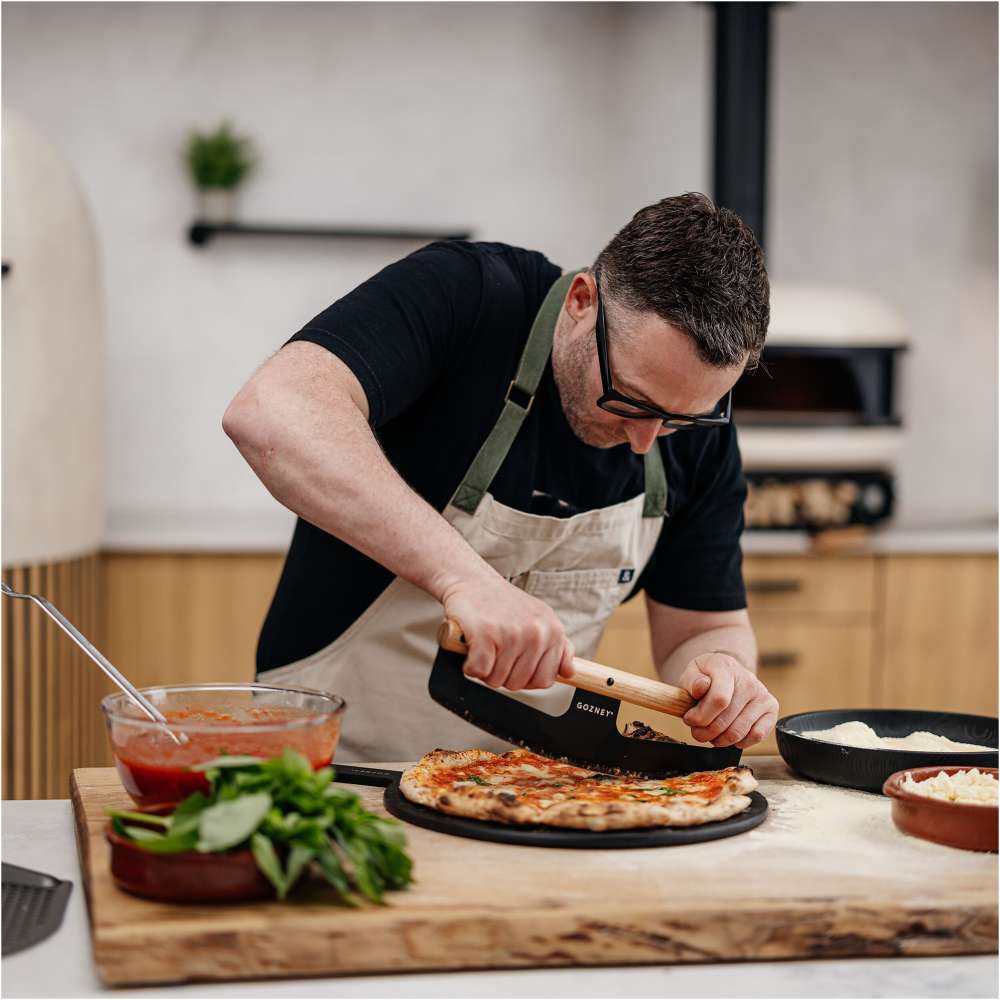Gozney Pizza Rocker being used by a man in an apron and glasses to cut a pizza on a wooden board in a kitchen with bowls of sauce basil and mozzarella nearby.