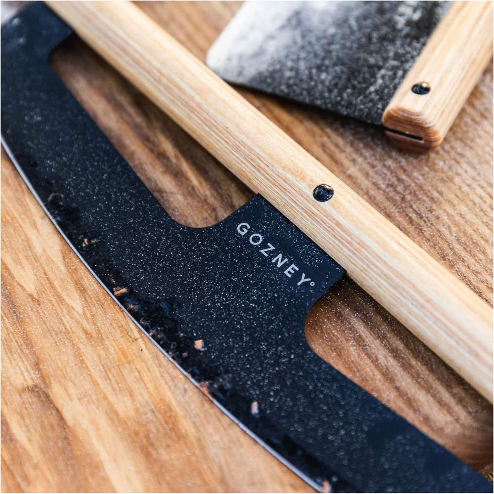 Gozney Pizza Rocker close view of the black curved blade and light wood handle resting on a floured wooden board with the Gozney wordmark on the blade.