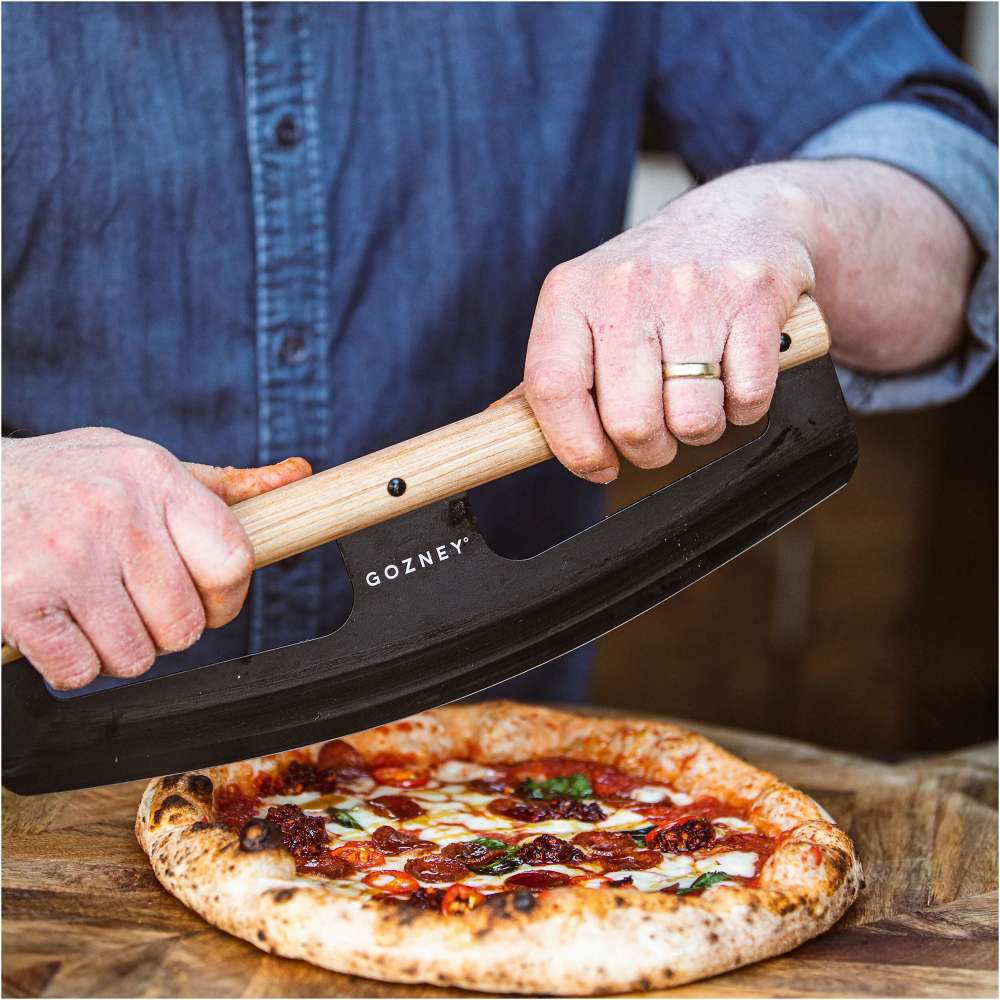 Gozney Pizza Rocker gripped in two hands by the wooden handle as it cuts through a baked pizza with the Gozney wordmark on the black blade.
