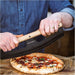 Gozney Pizza Rocker gripped in two hands by the wooden handle as it cuts through a baked pizza with the Gozney wordmark on the black blade.