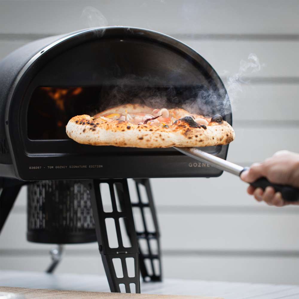 Gozney Roccbox Black Outdoor Portable Pizza Oven with a hand drawing a finished pizza out of the glowing mouth on a wooden peel and steam rising against a gray clapboard wall.