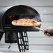 Gozney Roccbox Black Outdoor Portable Pizza Oven with a hand drawing a finished pizza out of the glowing mouth on a wooden peel and steam rising against a gray clapboard wall.