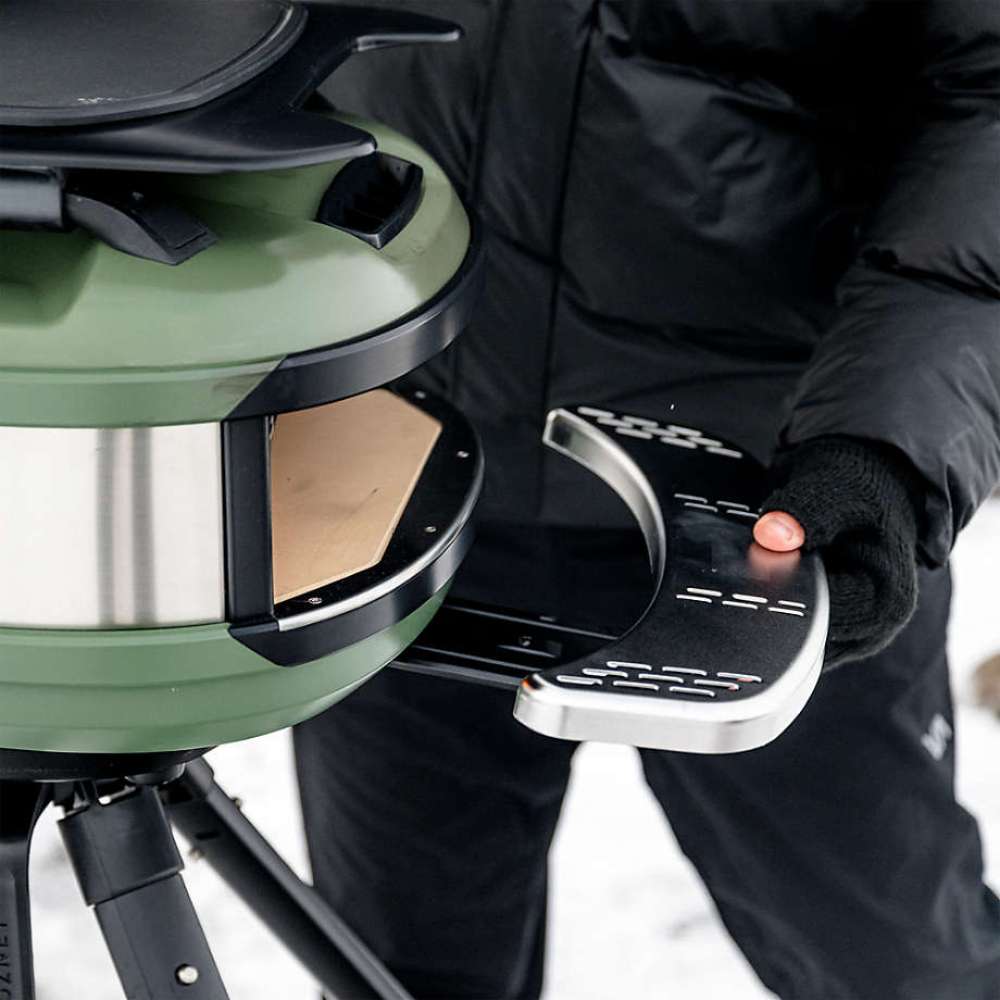 Gozney Tread Mantel being fitted to an Olive Tread oven on its tripod stand by a gloved hand in the snow.