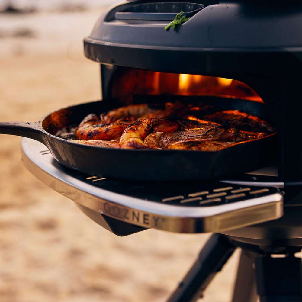 Gozney Tread Mantel holding a cast iron skillet of roast chicken at the mouth of a black Tread oven with sand behind.