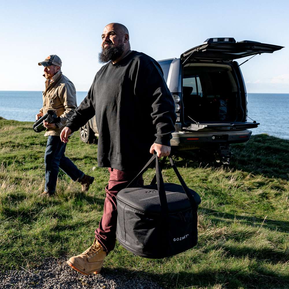 Gozney Tread Travel Bag carried by a bearded man across grass away from an open car tailgate with the sea and blue sky behind.