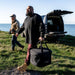 Gozney Tread Travel Bag carried by a bearded man across grass away from an open car tailgate with the sea and blue sky behind.