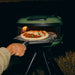 Gozney Venture Placement Peel at night launching a topped pizza into the glowing mouth of an Olive Tread oven on its tripod stand.