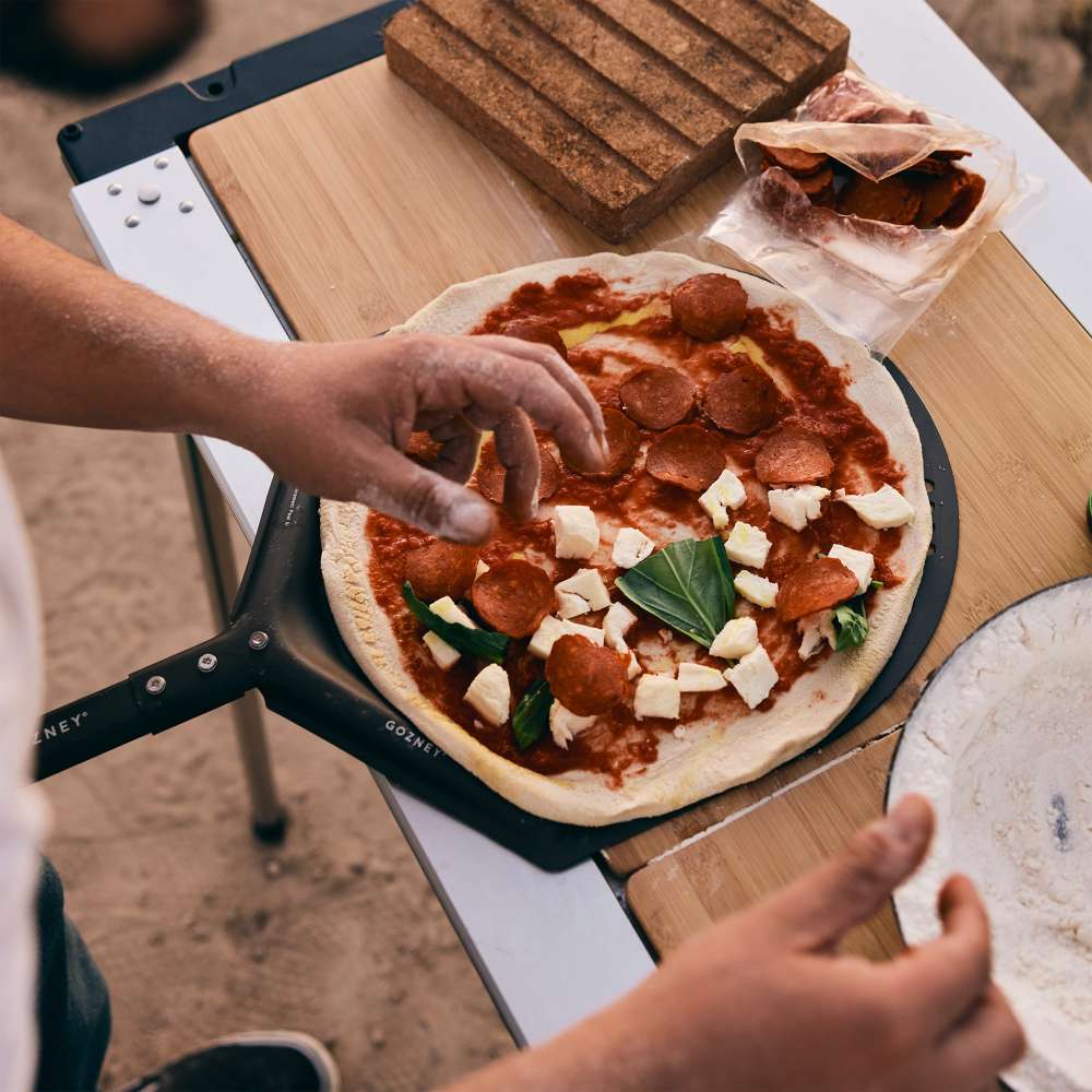 Gozney Venture Placement Peel overhead outdoors with hands topping a raw pizza with pepperoni and mozzarella on the perforated head resting on a folding table.