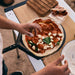 Gozney Venture Placement Peel overhead outdoors with hands topping a raw pizza with pepperoni and mozzarella on the perforated head resting on a folding table.