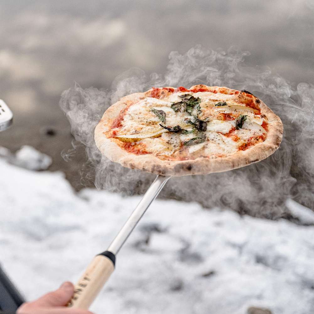 Gozney Venture Turning Peel held up with a finished margherita pizza on the head and steam rising against snowy ground.