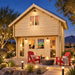 Illuminated Grand Loft 107 Deluxe DD in a landscaped Nevada backyard at night, with family and friends watching a sports game inside. Red patio chairs, a fire feature, pathway lights, desert plants, and mountains complete the scene.
