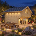 Two-story light wood loft with a dark gabled roof, glass doors, and a lit interior next to an outdoor wooden dining table.