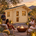 Front-left view of the Harvest Grand Bungalow 107 at golden hour. Four adults socialize around a fire feature while the compact illuminated backyard bar is visible through the studio’s door and windows.
