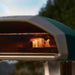 Highland Green Ooni Koda 2 Pizza Oven close view of the oven mouth with a flame inside against a blurred outdoor background.
