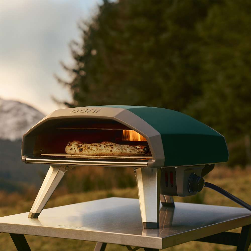 Highland Green Ooni Koda 2 Pizza Oven on a stainless table in a field at dusk with a cooked pizza on the glowing stone.