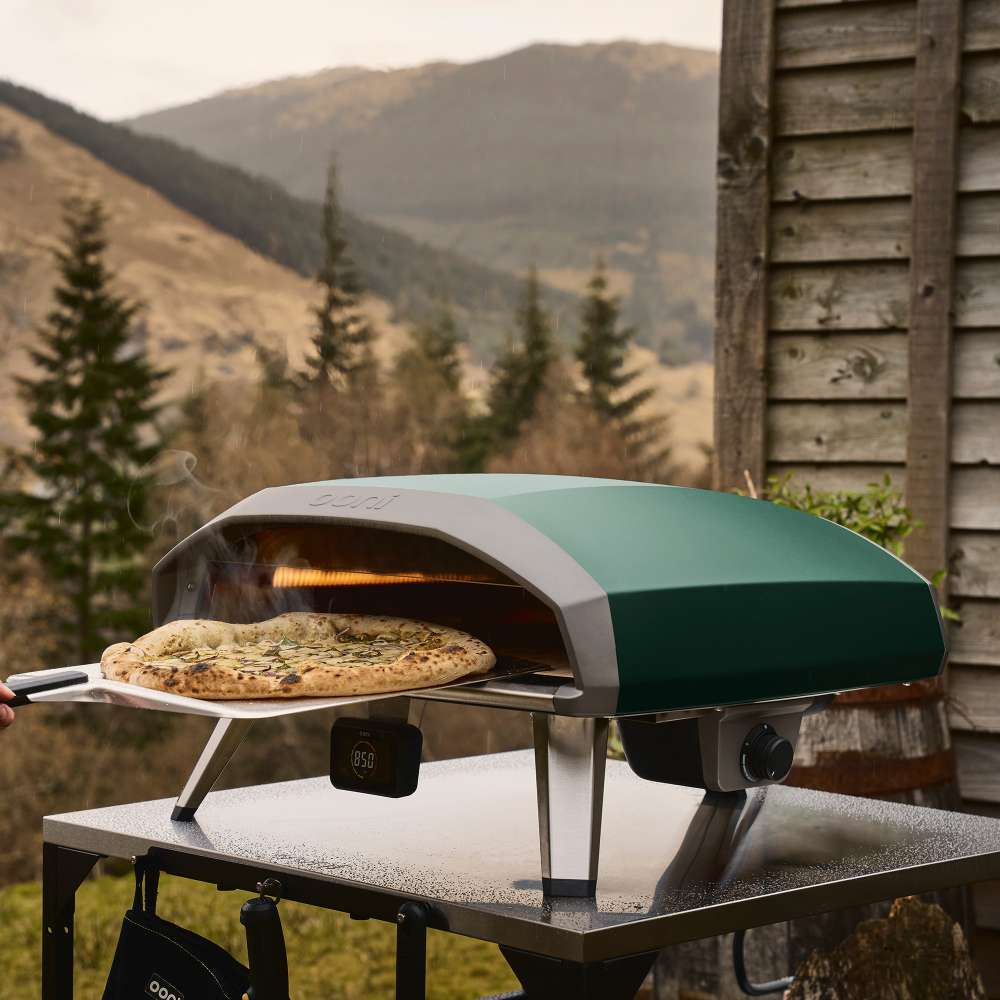 Highland Green Ooni Koda 2 Pro Pizza Oven on an outdoor table in a mountain valley with a cooked pizza on a peel at the mouth.