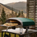 Highland Green Ooni Koda 2 Pro Pizza Oven on an outdoor table in a mountain valley with a cooked pizza on a peel at the mouth.