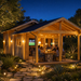 Nighttime exterior view of the illuminated Lake House 70A wooden cabin, showing people dining under string lights on the covered porch with a stone table and a stone pathway in the foreground.
