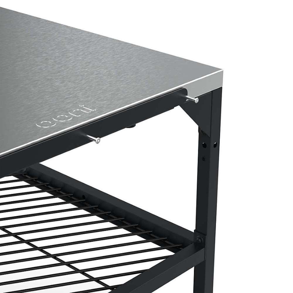 Large Ooni Modular Table close view of one corner of the stainless top with the ooni logo embossed and the mesh shelf below.