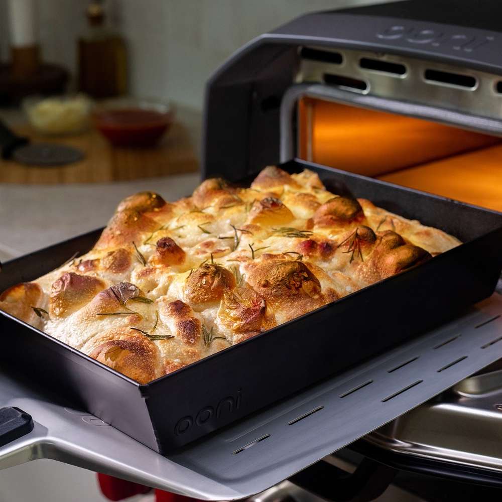 Medium Ooni Detroit Pizza Pan filled with a golden focaccia studded with garlic cloves and rosemary, resting on the ledge of an Ooni oven with its orange glow behind.