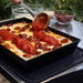 Medium Ooni Detroit Pizza Pan holding a baked pepperoni pizza while a hand spoons stripes of red sauce over the melted cheese on an outdoor counter.