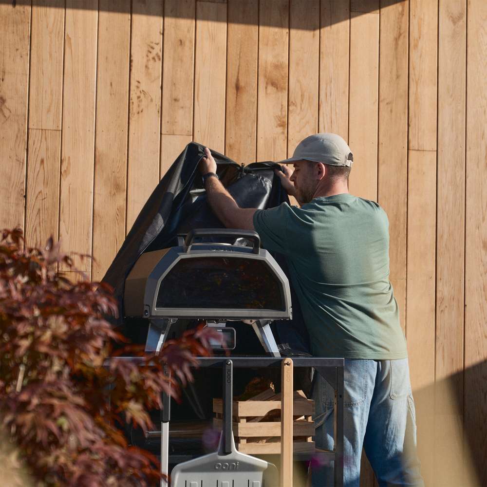 Medium Ooni Table and Pizza Oven Cover being pulled down over an oven on a table by a man in a green shirt against a timber wall.