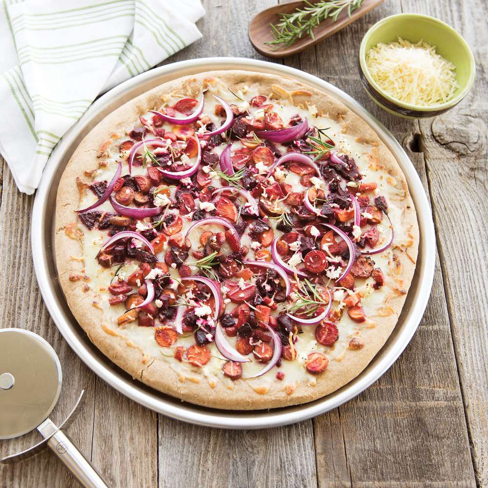 Nordic Ware Naturals 14" Deep Dish Pizza Pan on a weathered wood table holding a baked pizza with red onion tomato and olives beside a bowl of grated cheese and a pizza cutter.