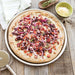 Nordic Ware Naturals 14" Deep Dish Pizza Pan on a weathered wood table holding a baked pizza with red onion tomato and olives beside a bowl of grated cheese and a pizza cutter.