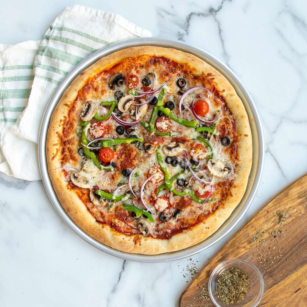 Nordic Ware Naturals 14" Traditional Pizza Pan close from above with a baked pepper olive and onion pizza in it next to a wooden board and a bowl of dried herbs.