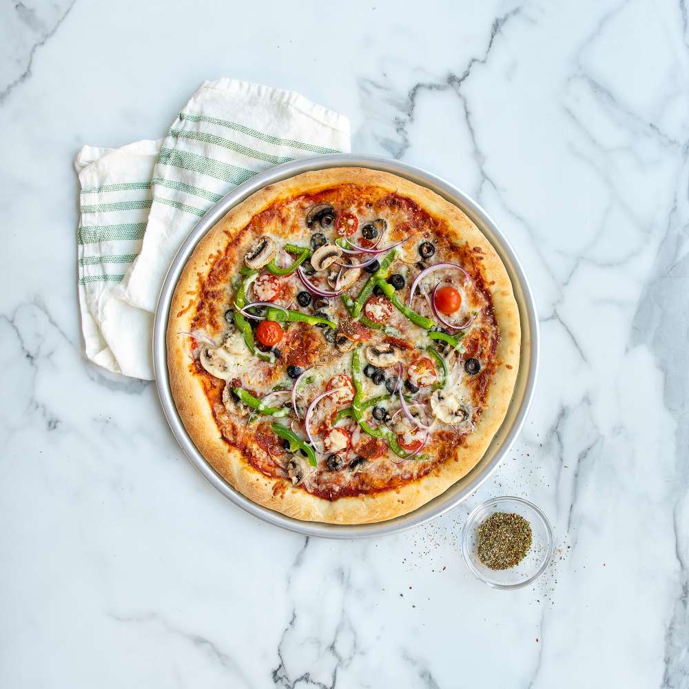 Nordic Ware Naturals 14" Traditional Pizza Pan from above on pale marble holding a baked vegetable pizza beside a striped towel and a small bowl of dried herbs.
