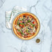 Nordic Ware Naturals 14" Traditional Pizza Pan from above on pale marble holding a baked vegetable pizza beside a striped towel and a small bowl of dried herbs.