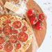 Nordic Ware Naturals 9" Air Crisper Pizza Pan lifestyle shot of a baked tomato and herb pizza resting on a round wooden board beside vine tomatoes and a wedge of parmesan.