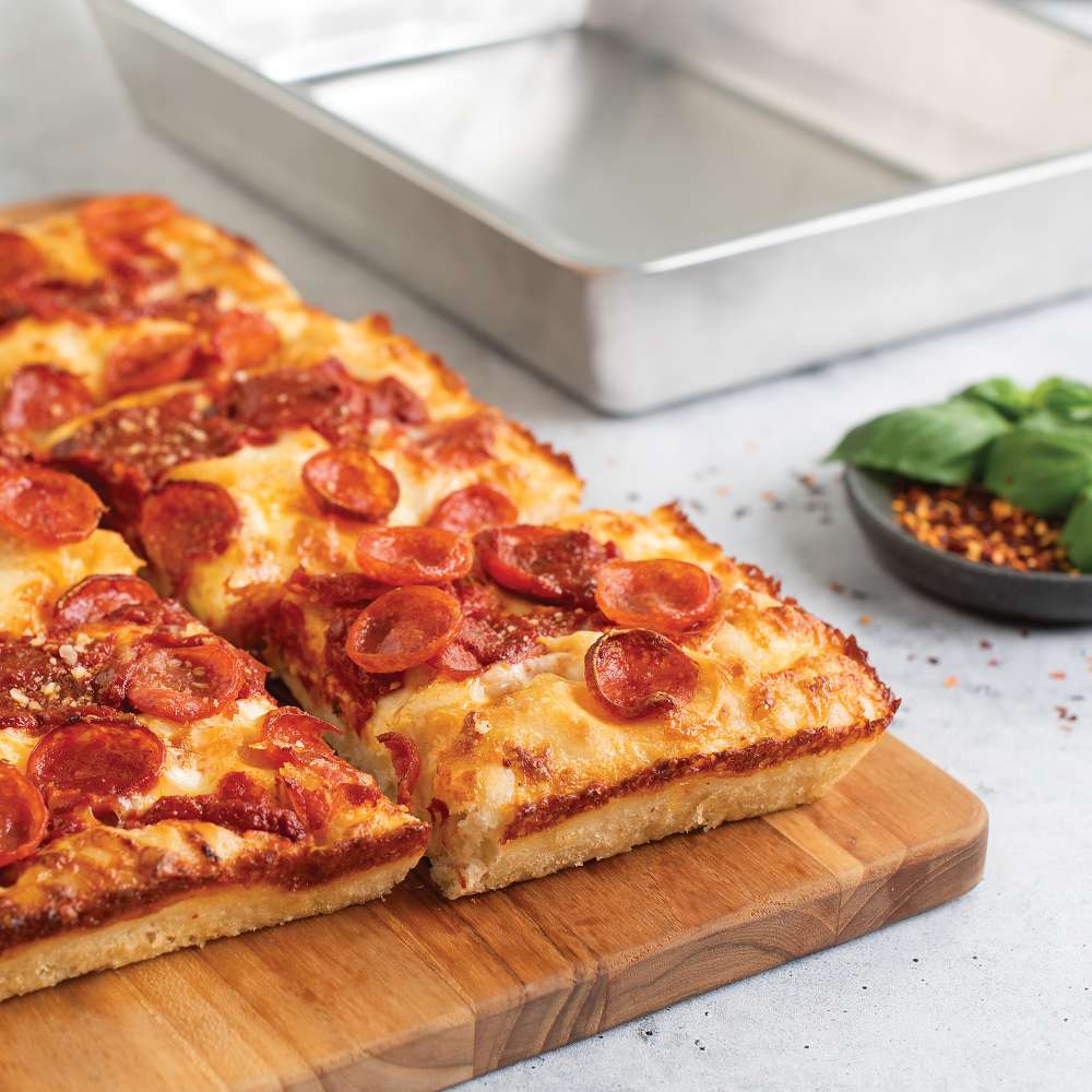 Nordic Ware Naturals Detroit-Style Pizza Pan behind a sliced pepperoni pizza on a wooden board with a dish of basil and chili flakes.