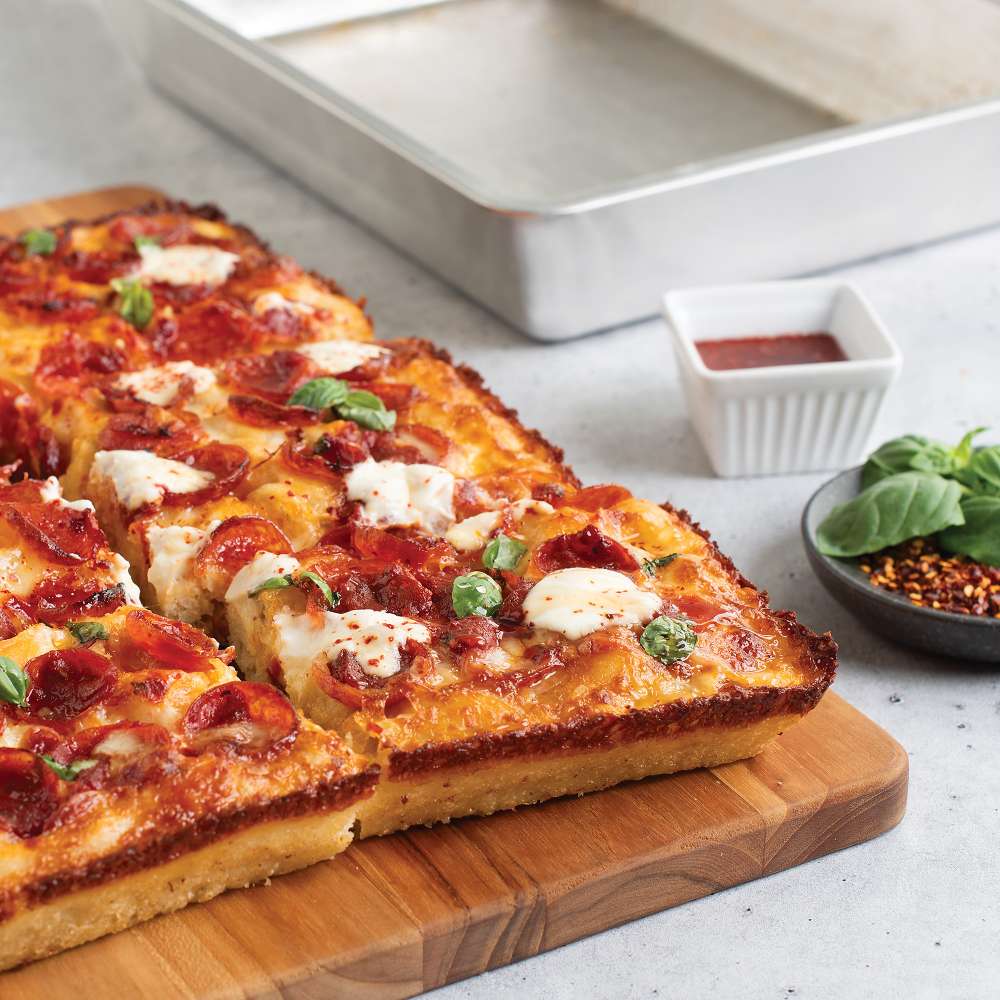 Nordic Ware Naturals Detroit-Style Pizza Pan behind a sliced pizza topped with pepperoni mozzarella and basil on a wooden board with a ramekin of sauce.