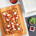 Nordic Ware Naturals Detroit-Style Pizza Pan from above beside a whole pepperoni and mozzarella pizza on a wooden board with bowls of sauce basil and chili flakes.