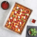 Nordic Ware Naturals Detroit-Style Pizza Pan from above holding a baked pepperoni mozzarella and basil pizza with a bowl of sauce and a dish of basil and chili flakes beside it.