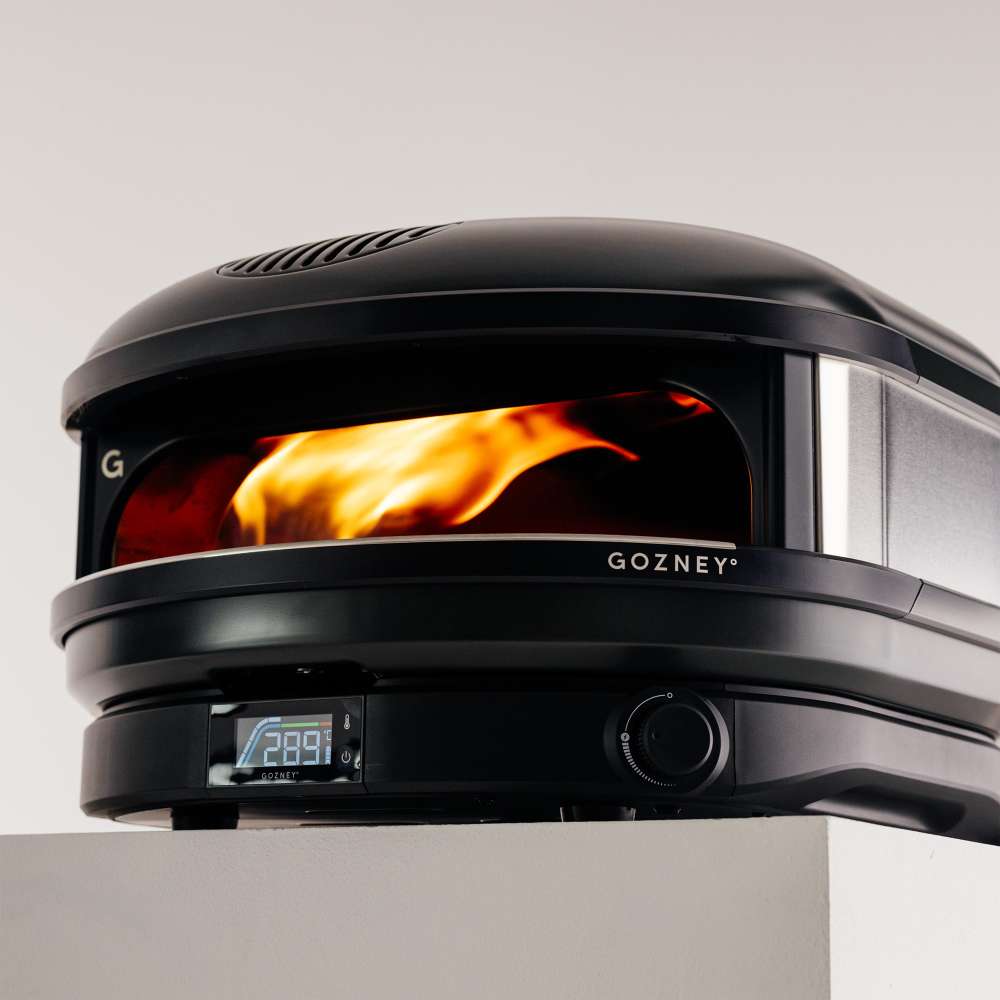 Off-Black Gozney Arc XL Pizza Oven close view on a pale countertop with a tall orange flame filling the copper-toned dome and the control dial in the foreground.