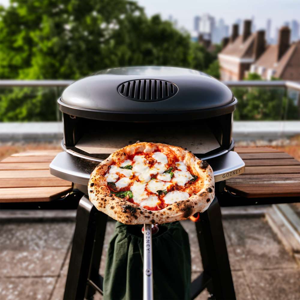 Off-Black Gozney Arc XL Pizza Oven on a black stand on a rooftop terrace with a leopard-spotted margherita pizza held on a peel in front of it and trees and rooftops behind.