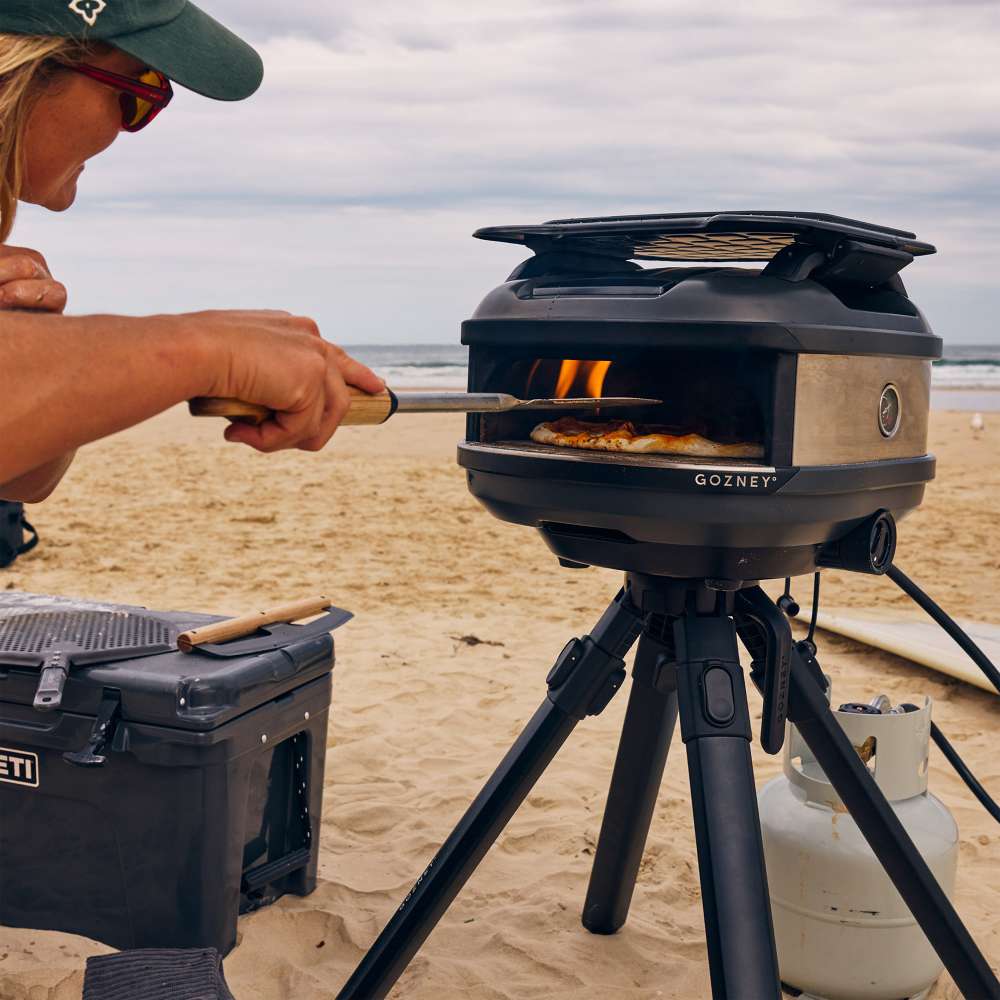 Off-Black Gozney Tread Pizza Oven on its tripod stand at the beach with a person in a cap sliding a pizza in on a peel and a gas cylinder and cooler alongside.