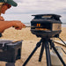 Off-Black Gozney Tread Pizza Oven on its tripod stand at the beach with a person in a cap sliding a pizza in on a peel and a gas cylinder and cooler alongside.