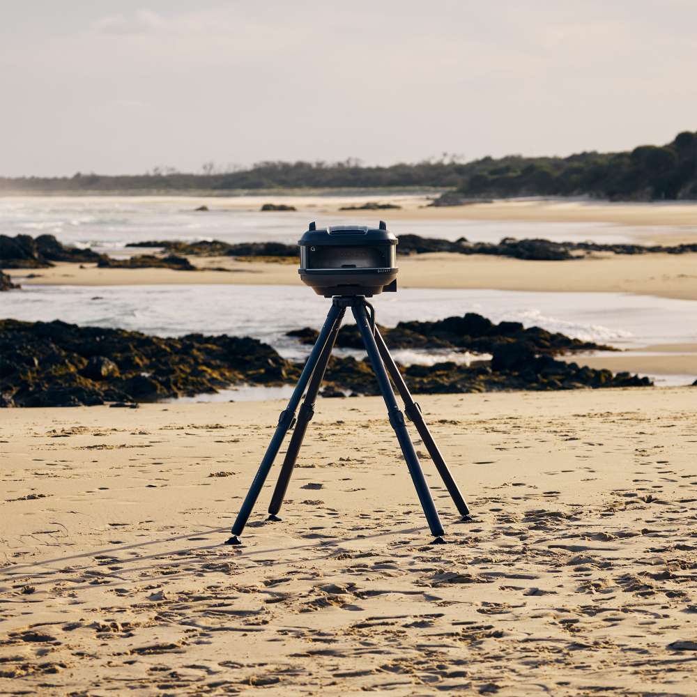 Off-Black Gozney Tread Pizza Oven set up on its folding tripod stand on an empty sand beach with rocks and the sea behind.
