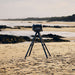 Off-Black Gozney Tread Pizza Oven set up on its folding tripod stand on an empty sand beach with rocks and the sea behind.