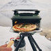 Olive Gozney Tread Pizza Oven on its tripod stand in a snowy mountain landscape with a cooked pizza at the mouth and a hand holding a wooden peel.
