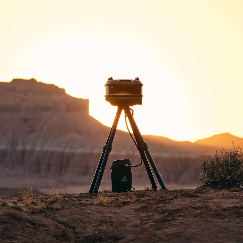Olive Gozney Tread Pizza Oven silhouetted on its tripod stand at sunset in a desert landscape with red rock cliffs behind and a gas cylinder on the ground.