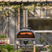 Ooni Karu 2 Pro Pizza Oven on a garden table under a leafy pergola with flames glowing inside the oven mouth.