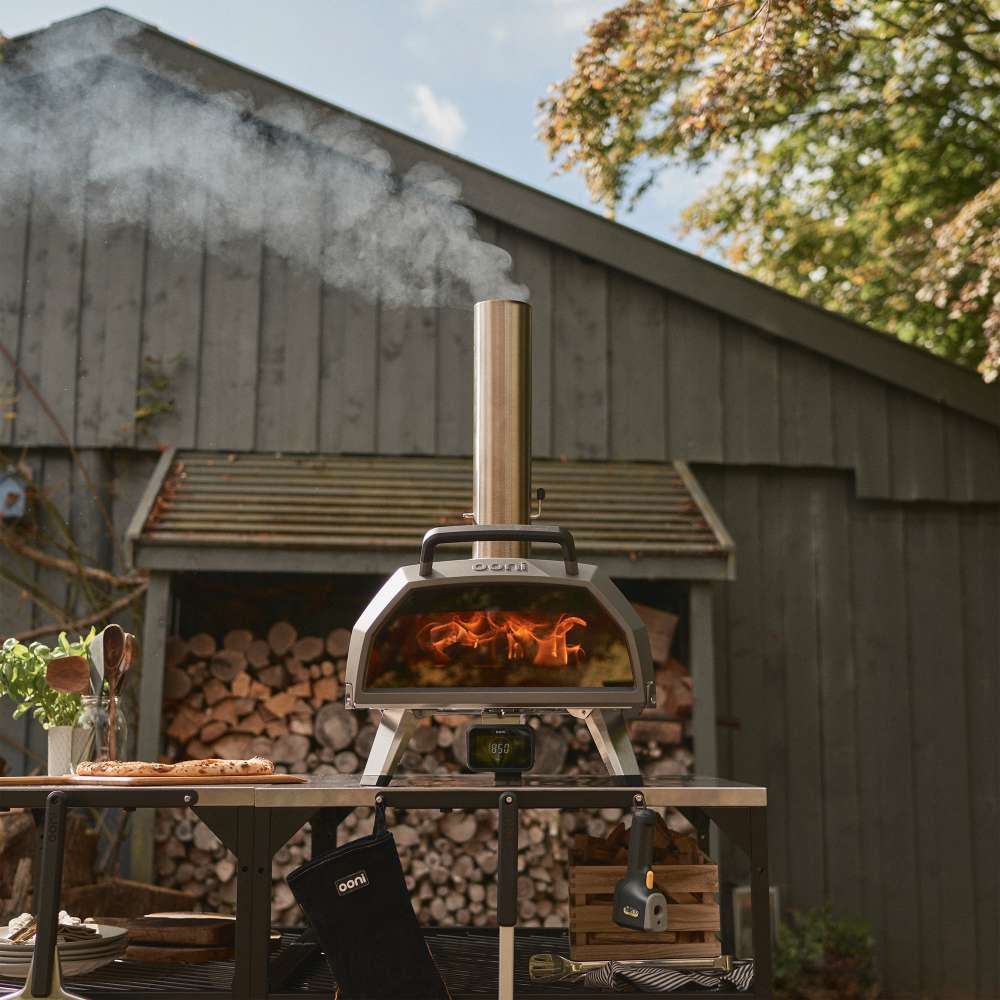Ooni Karu 2 Pro Pizza Oven on an outdoor counter beside a grey timber building with smoke rising from the chimney and firewood stacked behind.