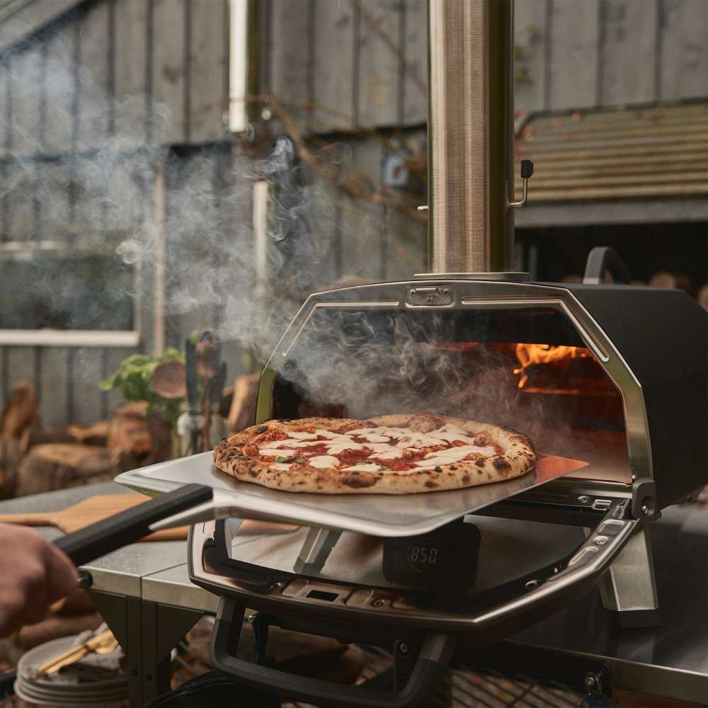 Ooni Karu 2 Pro Pizza Oven with a hand sliding a topped pizza on a peel into the fiery mouth as smoke rises.
