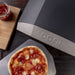 Ooni Koda 12 Outdoor Pizza Oven close view of the grey shell corner with the ooni wordmark beside a cooked pizza on a plate.