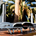 Ooni Koda 12 Outdoor Pizza Oven lined up with other lit Ooni ovens on a deck with palm trees behind.