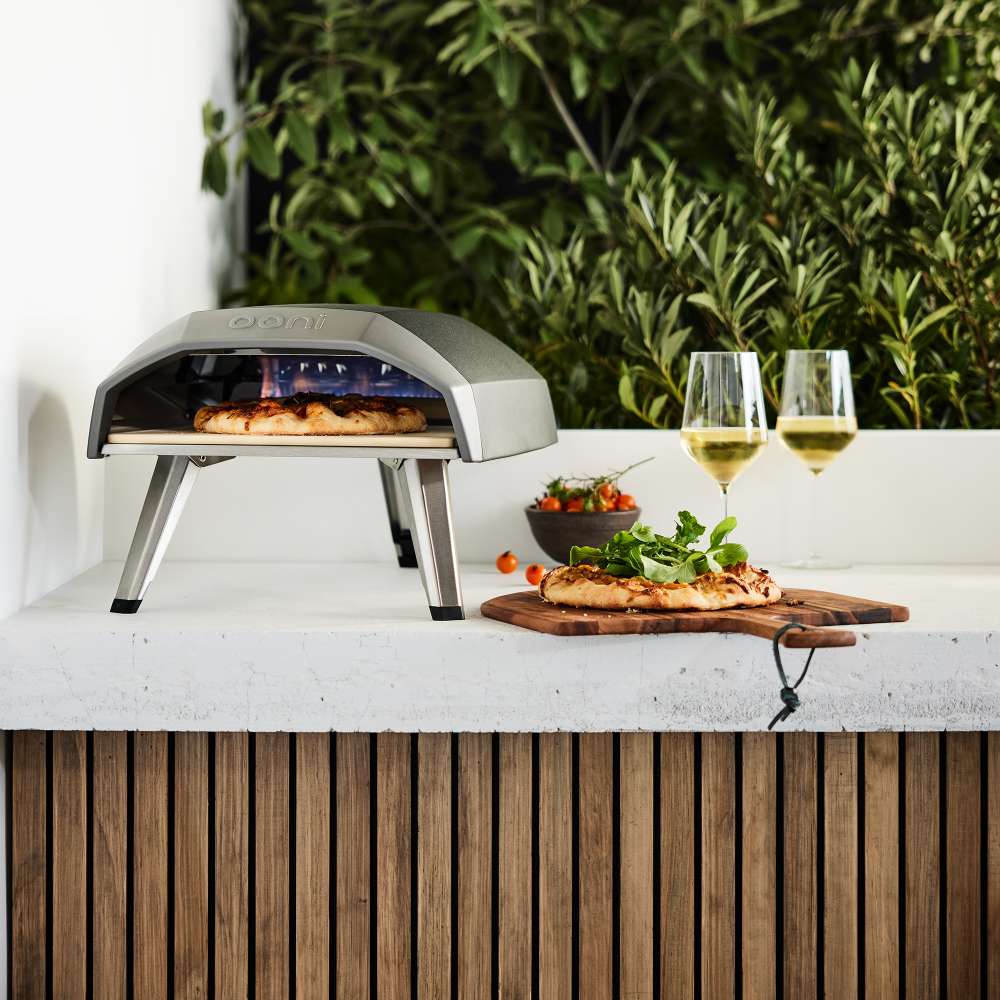 Ooni Koda 12 Outdoor Pizza Oven on a pale concrete counter with a cooked pizza on a board, two glasses of white wine and greenery behind.